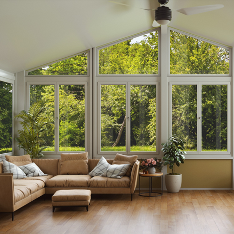 The interior of a sunroom with tall windows and a peaked ceiling with a tan L-shaped couch with an ottoman in front of it and plants next to it and trees outside the windows.  